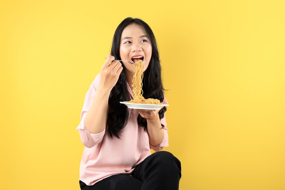 Young Asian Woman Holding A Plate of Fried Instant Noodle, Sitting on Yellow Background