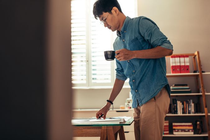 Side view of man with coffee standing at table looking at building plan