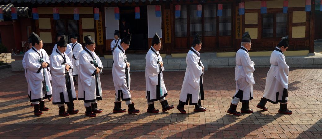 Korean men in light robe and dark hat walking in line beside building