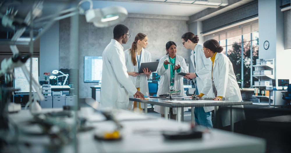 Group of International Multiethnic Lab Specialists Gathering Around a Table to Discuss Blueprints and Components for a High-Tech Robotic Project