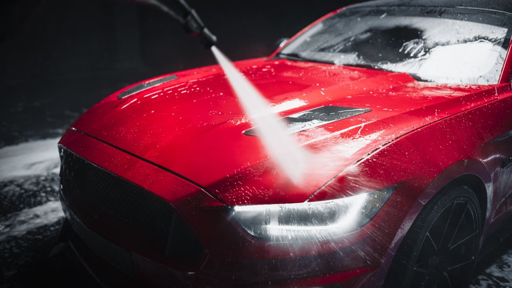 Car Wash Expert Using Water Pressure Washer to Rinse a Red Modern Sportscar.