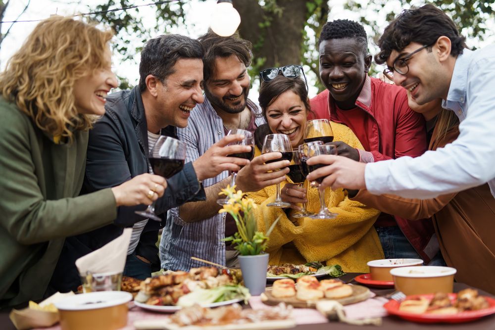Adults around a set table, toasting with red wine, under decorative lights and trees