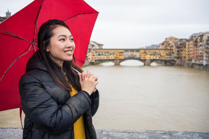 Cheerful Asian tourist with umbrella standing on bridge near river
