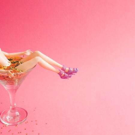 Doll bathing in martini glass full of gold glitter on pink background