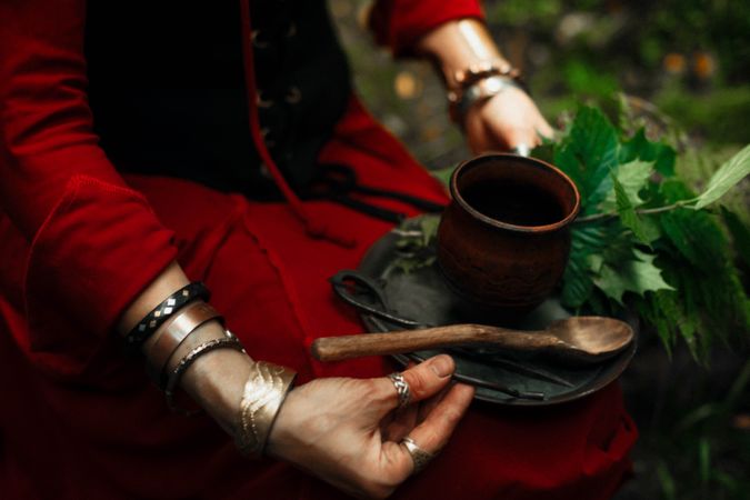 Cropped image of witch holding plate of herbs with spoon and mug