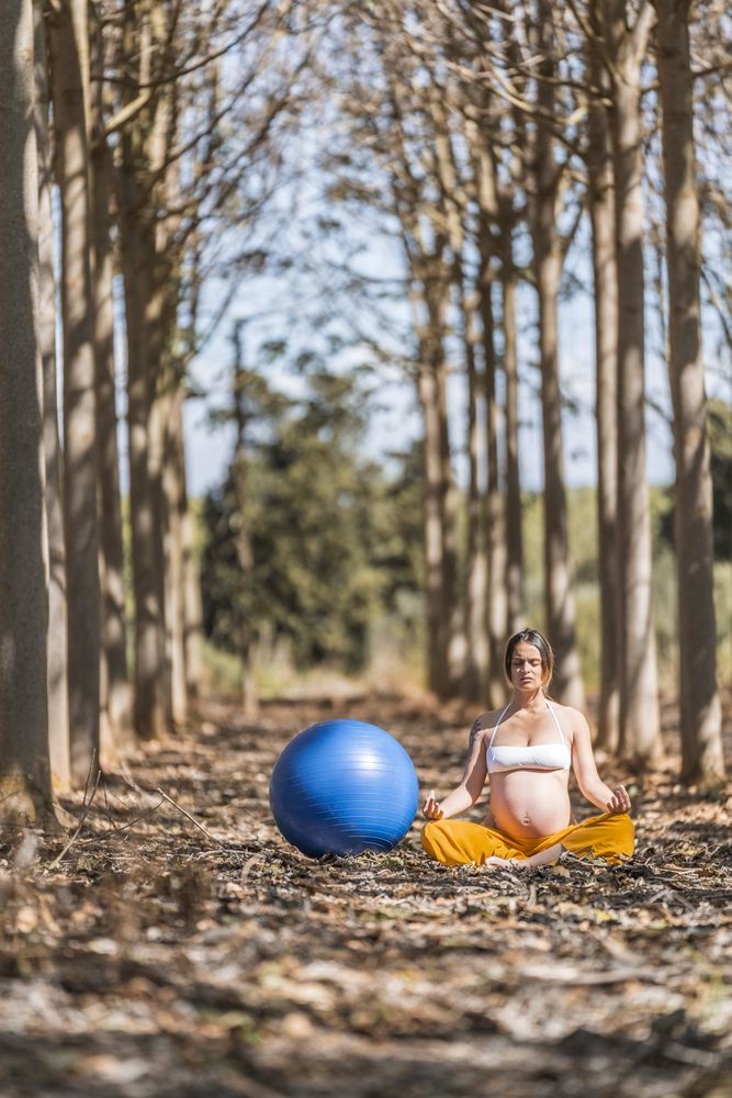 Serene adult pregnant woman meditating while sitting on ground among trees in park during sunny day