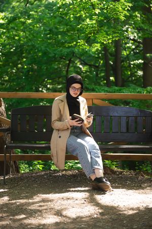 Woman in headscarf sitting on park bench on sunny day with book