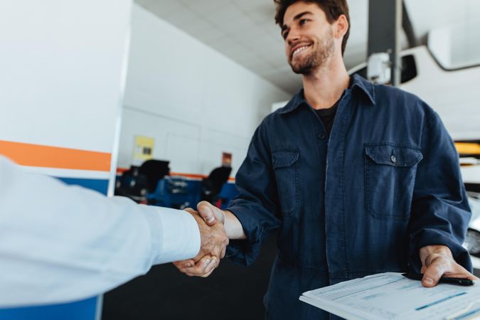 Young auto mechanic shaking hands with satisfied customer in garage