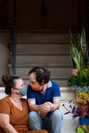 Loving partners look at each other smiling while sitting on stairs outside wearing masks
