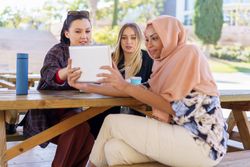 Three women on park bench with insulated mugs watching digital tablet
