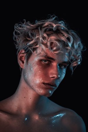 Portrait of topless blonde man with UV paint on his face against dark background