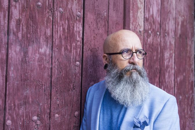 Front view of a mature trendy bearded hipster leaning on a vintage red door outdoors while looking away