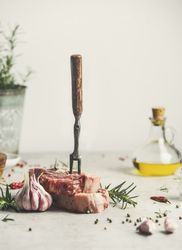 Raw meat steak with ingredients and meat fork on kitchen table with rosemary, pepper and olive oill life