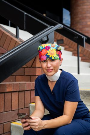 Woman nurse in floral head covering and scrubs sits during break with smart phone looking at camera