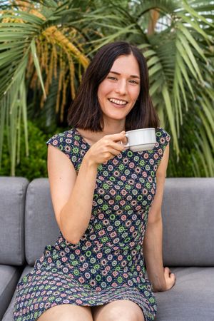 A smling woman is sitting on a couch and holding a cup of coffee