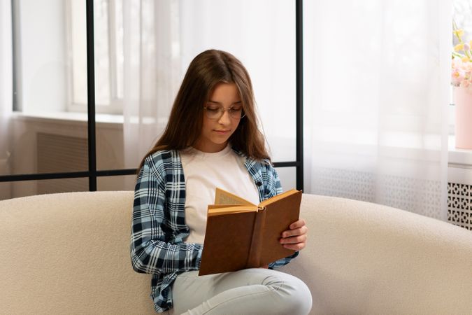 Girl wearing glasses sitting on sofa reading book calmly.