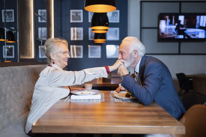 Grey haired man taking hand of woman in restaurant to kiss as she smiles