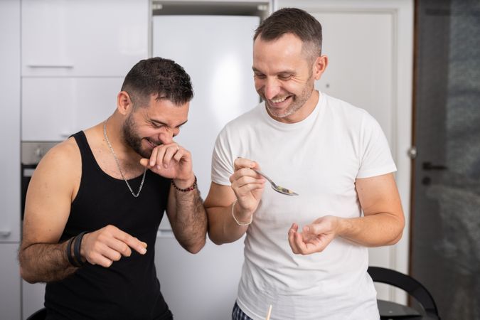 Laughing male couple tasting pasta as they prepare meal