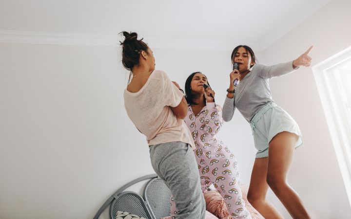 Young women having fun singing karaoke standing on bed