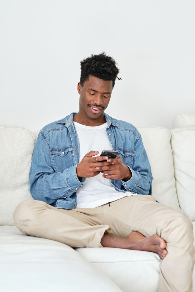 Smiling Black man browsing smartphone on cozy sofa