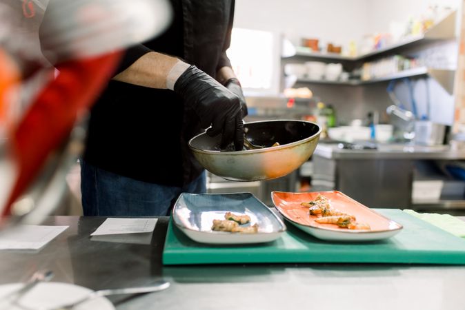 Side view of crop anonymous male cook in gloves standing at table and serving delicious broiled prawn with vegetables on plates in kitchen