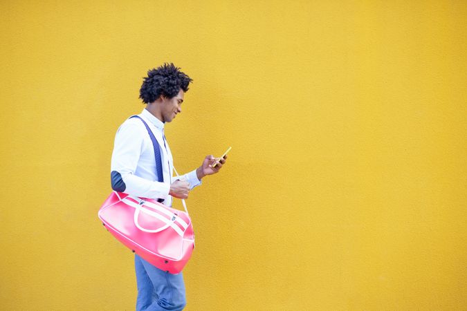 Man with bright pink back smiling at phone walking next to yellow wall