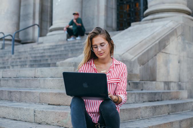 Chic woman sitting on outdoor steps in European city working on her laptop