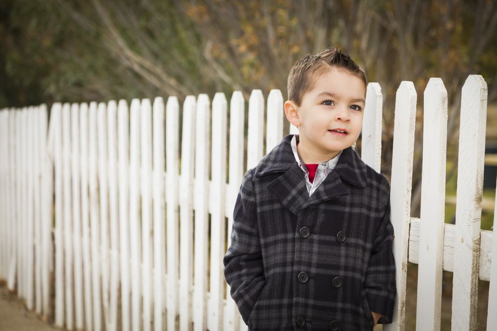 Young Boy Waiting For School Bus Along Fence Outside.