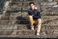 Man in woolen sweater sitting on steps outside thinking with shadows of trees beVB30