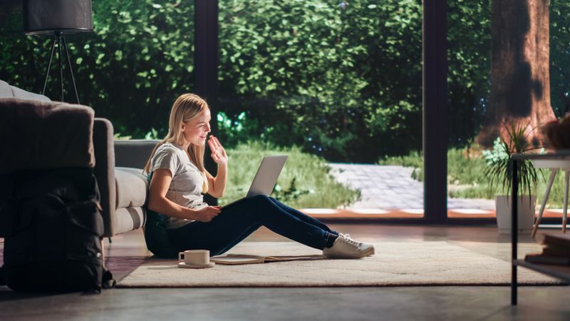 Portrait of Beautiful Young Adult Female with Blond Hair Sitting on a Floor and Using Laptop Computer to Make a Video Call in Sunny Living Room.