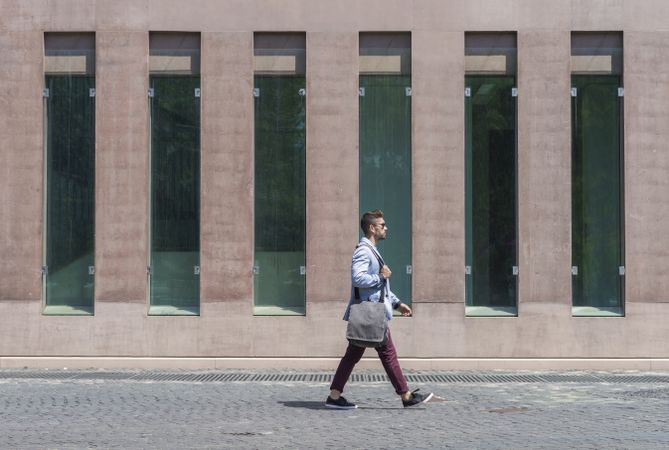 Side view of young businessman walking next to office buildings while holding a shoulder bag outdoors