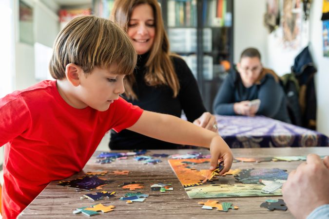 Concentrated preschooler playing puzzle game at with crop parents