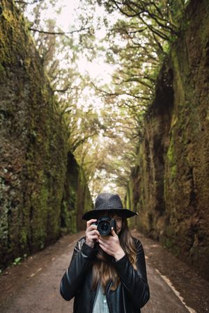 Young stylish woman taking photo on camera on way between high walls and trees
