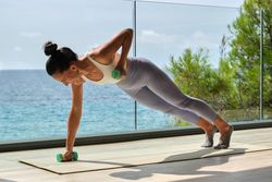 Sportswoman doing plank and raising arms with dumbbells in sunlight