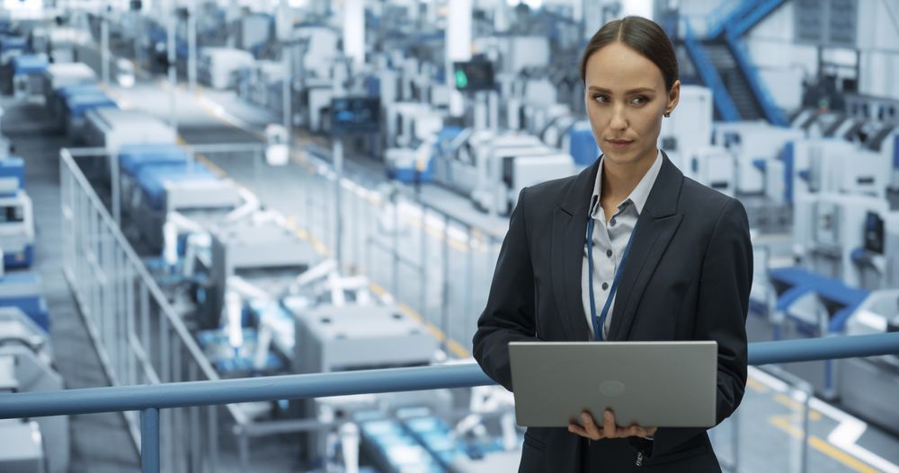 Portrait of a Female Engineer Using Laptop Computer and Looking Around a Factory Facility with Robotic Arms Producing Modern Electronic Components for Tech Industries