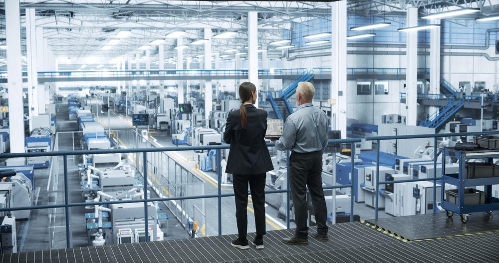 Mature Engineer and Female Scientist Standing with Their Back, Using Laptop Computer and Talking in a Factory Facility with Equipment Producing Modern Technical Devices for Different Industries