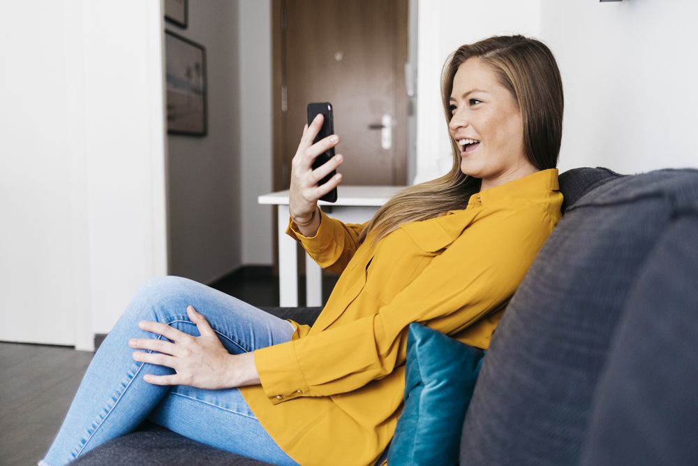 Beautiful adult female sitting on sofa while taking videocall on smartphone