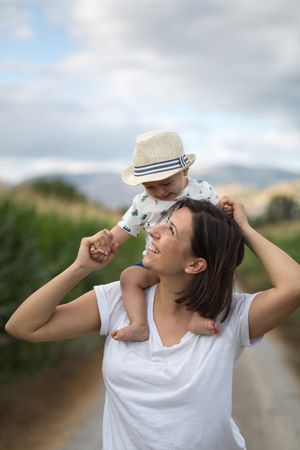 Adorable kid in hat on shoulders and looking at mother