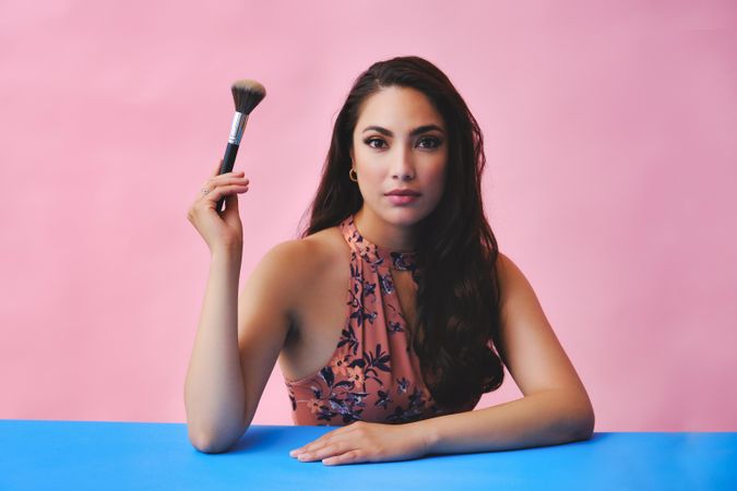 Hispanic woman with long brown hair holding large make up brush up and looking at camera