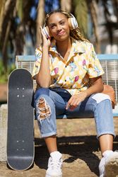 Happy female in bold patterned shirt sitting on park bench listening to music on large headphones