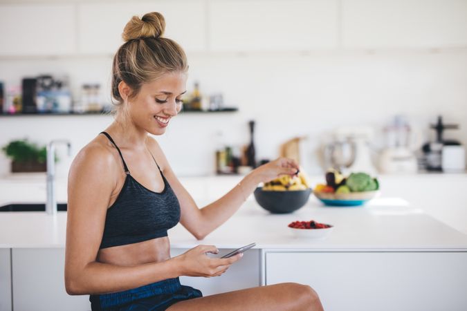 Smiling young woman sitting in kitchen using mobile phone and eating fruits