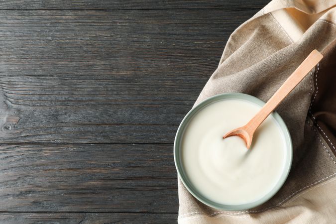 Bowl of sour cream yogurt, spoon and towel on wooden background