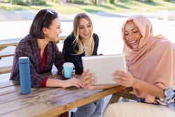 Three women on park bench with insulated mugs looking at something on digital tablet