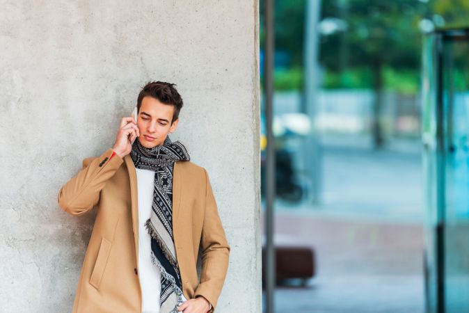 Man leaning on wall outside speaking on phone and wearing a camel coat 