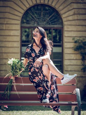 Cheerful dreamy woman sitting on back of bench with flowers