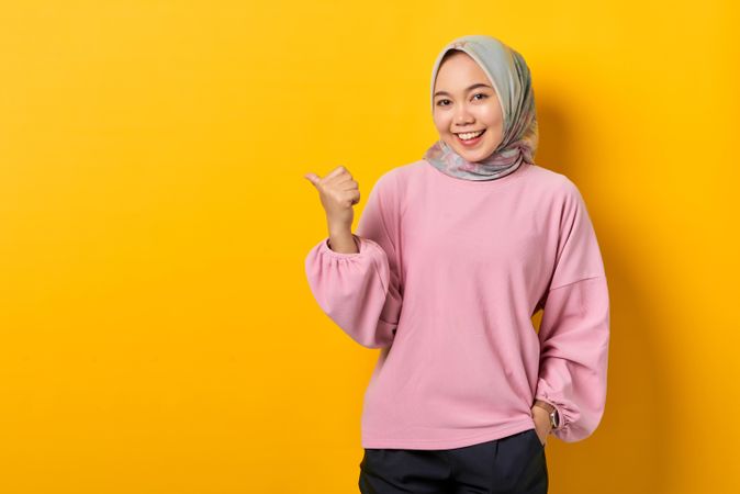 Smiling Muslim woman in scarf and pink sweater pointing to side with thumb