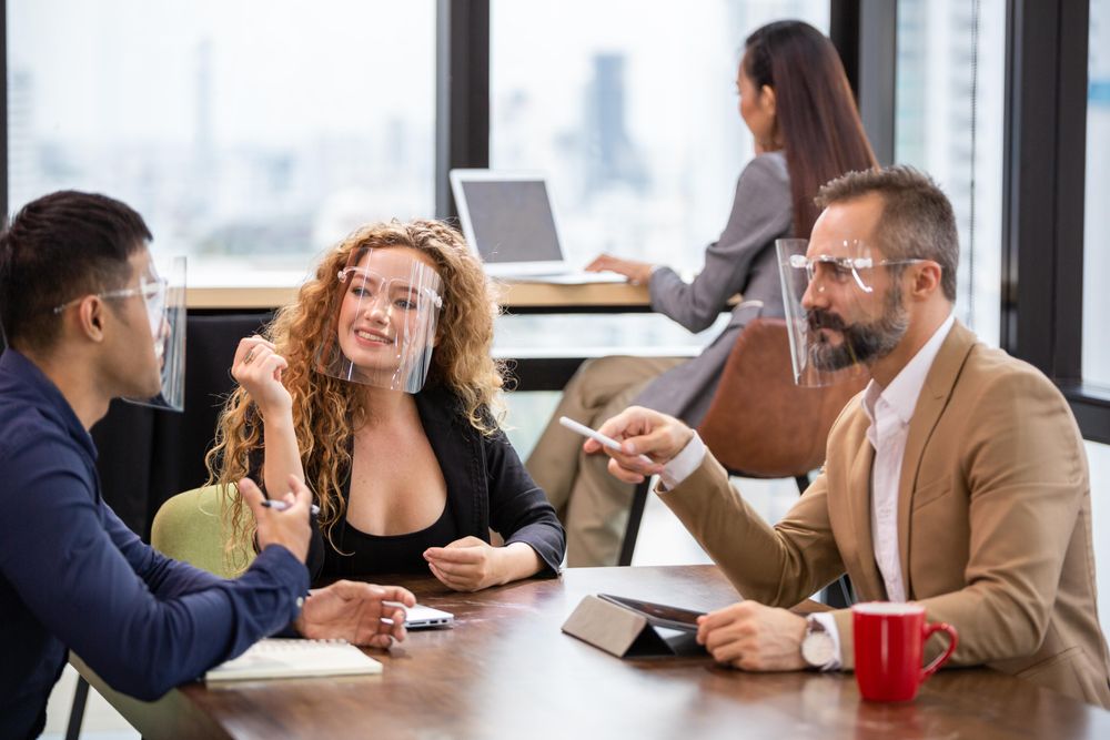 Happy colleagues discussing work in co-working space while wearing clear face shields
