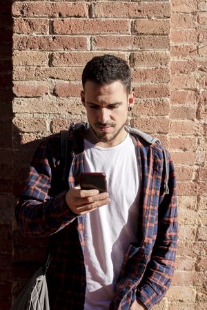 Young bearded male leaning on a brick wall checking his smartphone