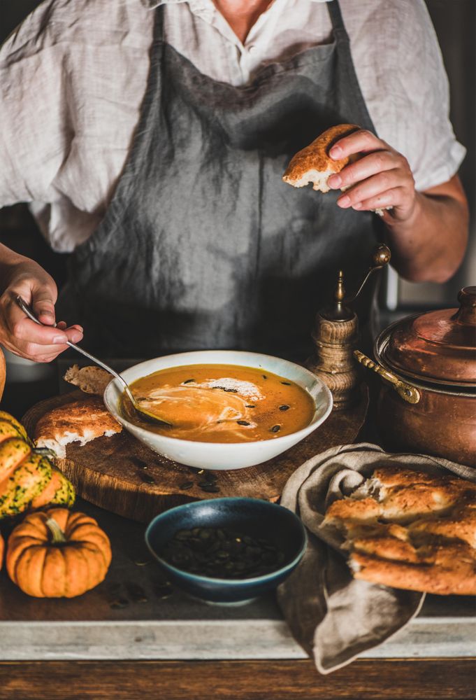 Woman in apron with spoon and bread eating soup in cozy kitchen