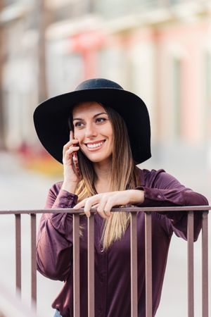 Charming woman in trendy outfit talking on smartphone and smiling at city street